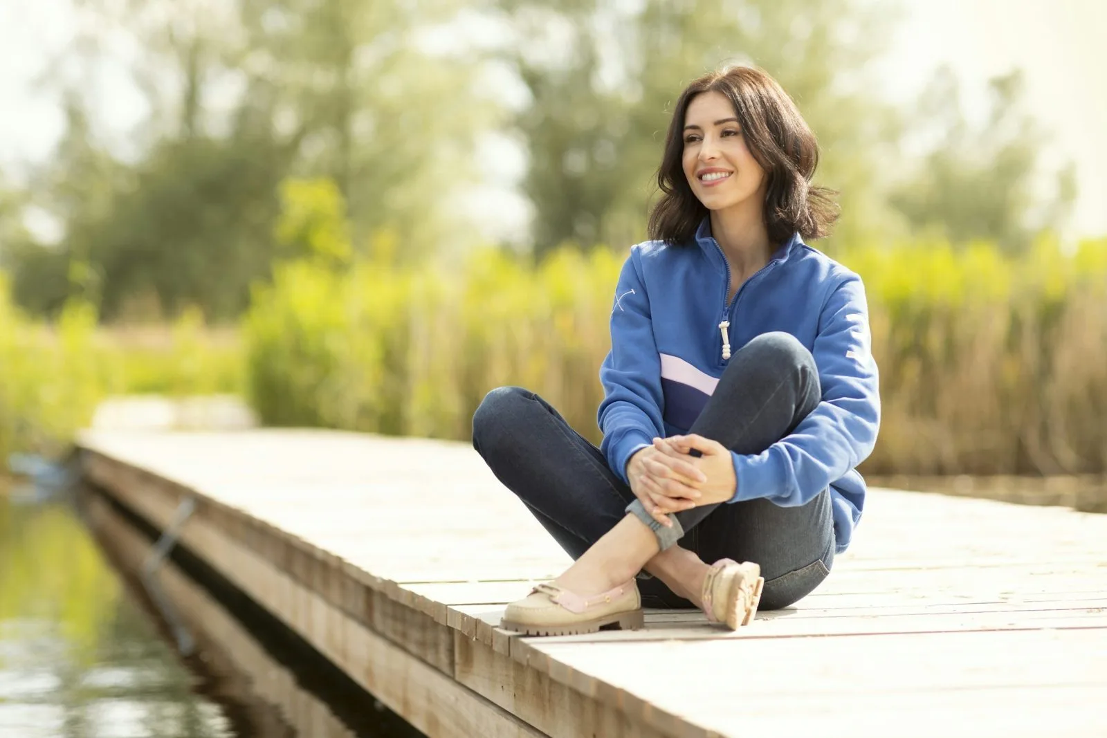 Woman in blue scrubs smiling while sitting on a wooden dock near water and trees